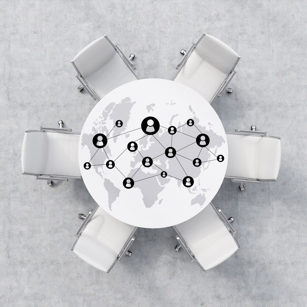 Top-down view of a white circular conference table surrounded by six office chairs on a concrete floor. The tabletop features a world map overlaid with a complex network diagram of interconnected person icons, symbolizing the 'Buying Group' and the shift from the single-buyer myth to account-based intelligence in B2B sales.